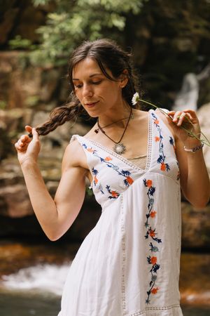Brunette woman in white embroidered sundress holding a daisy and her braid near a serene waterfall.