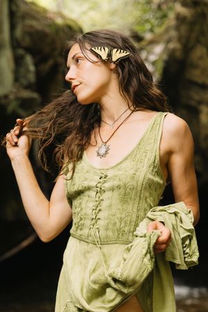 Woman in green dress with butterfly hair accessory poses in forest near rocks.