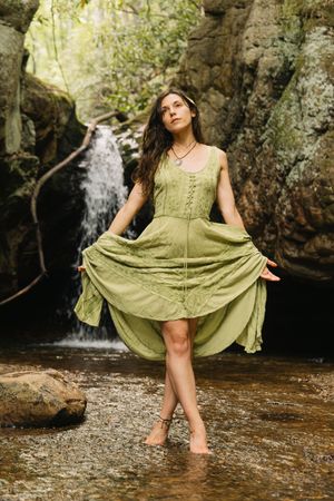 Woman in green dress standing barefoot in a stream near a waterfall in a lush forest setting.
