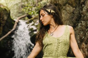 Woman in green dress with butterfly hair accessory meditating near a waterfall in a lush forest setting.