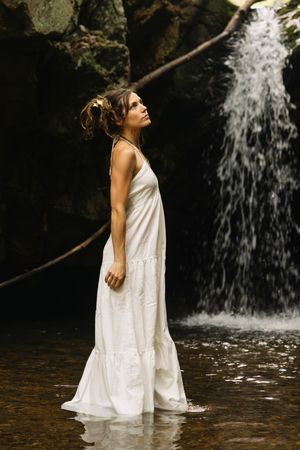 Woman in white maxi dress standing in shallow water before a serene waterfall in a rocky forest setting.