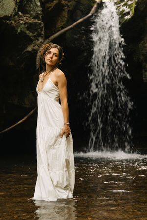 Woman in white maxi dress standing in water before a rocky waterfall in a lush forest setting.