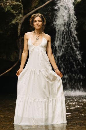 Woman in white tiered halter dress standing confidently in water before a lush forest waterfall.