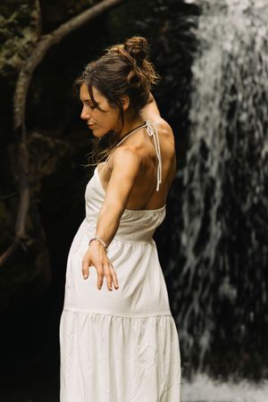 Woman in backless white halter dress posing gracefully in front of a serene waterfall.