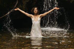 Woman in white dress stands in stream with arms raised near waterfall.