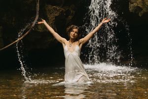 Woman in wet white dress holds branch with arms raised in waterfall pool.