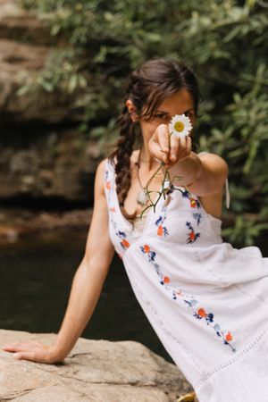 Young woman in white floral dress sitting on rocks, holding a daisy flower toward the camera outdoors.