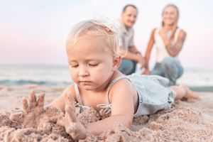 A baby girl plays in the sand at the beach while her parents smile in the background.