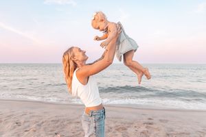 A mother joyfully lifts her baby girl in the air on a sandy beach at sunset with ocean waves in the background.