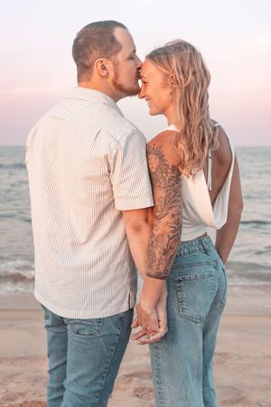 Romantic couple sharing a kiss on the beach at sunset, woman with tattoos holding hands in jeans.