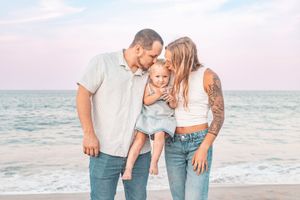 A happy family of three poses on a beach, parents kissing their baby girl with ocean waves in the background.