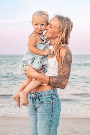 Tattooed mother holding her toddler daughter on a beach at sunset, both smiling and sharing a tender moment.