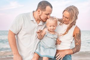 A young family with a tattooed mother and father kissing their baby girl on a sunny beach.