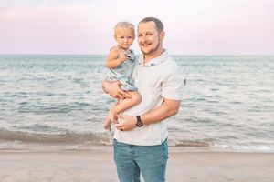 A smiling father holds his toddler on a sandy beach at sunset with calm ocean waves in the background.