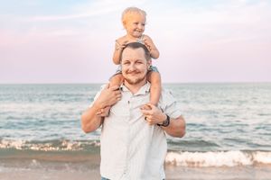 A smiling father carries his happy toddler on his shoulders at a beach during sunset.