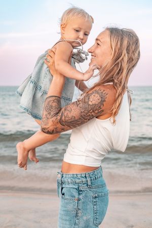 Tattooed mother joyfully lifting her toddler daughter at the beach, both smiling, wearing denim outfits near the ocean.