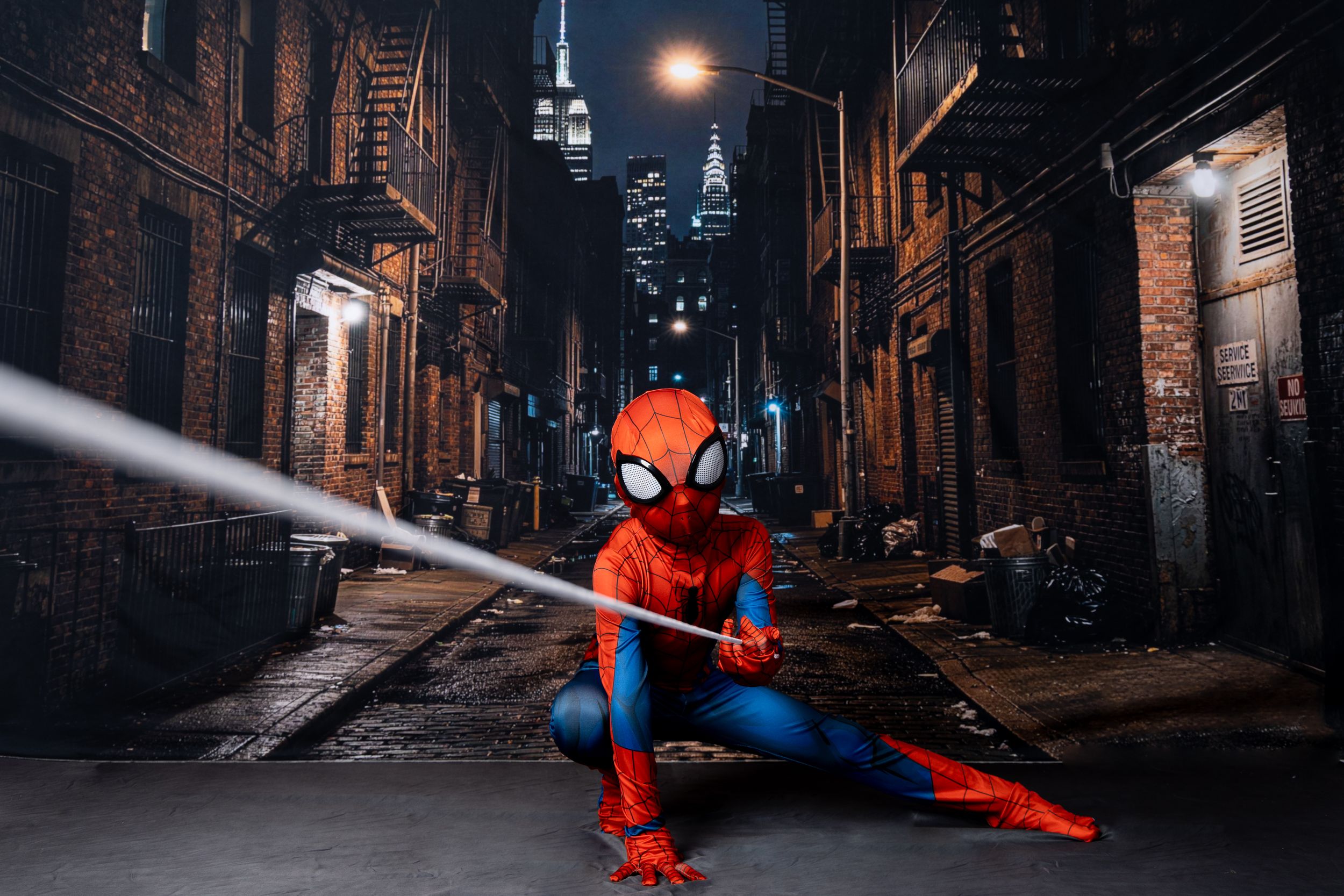 Spider-Man cosplayer crouches in a dark city alley shooting a web, with brick buildings and a glowing skyline behind.