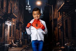 Smiling young boy in a Spider-Man costume opens his white shirt on a dark city alley set with fire escapes and a skyscraper behind.