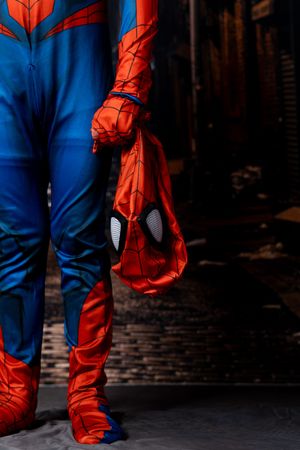 Person in a blue and red Spider-Man costume holding the unmasked red web-patterned mask beside their leg in a dark urban alley.