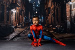 Young child in a red and blue Spider-Man costume crouching in a superhero pose in a dimly lit city alley with brick buildings and skyline.