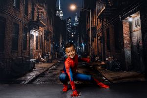 Young child in a red and blue Spider-Man costume crouching in a dark city alley at night with brick buildings and skyscrapers behind.
