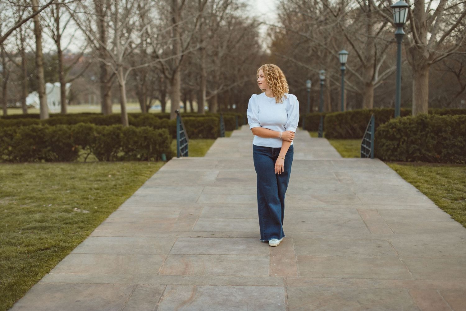 A senior girl stands in the middle of a sidewalk with ankles crossed, holding one arm and looking off to the side.