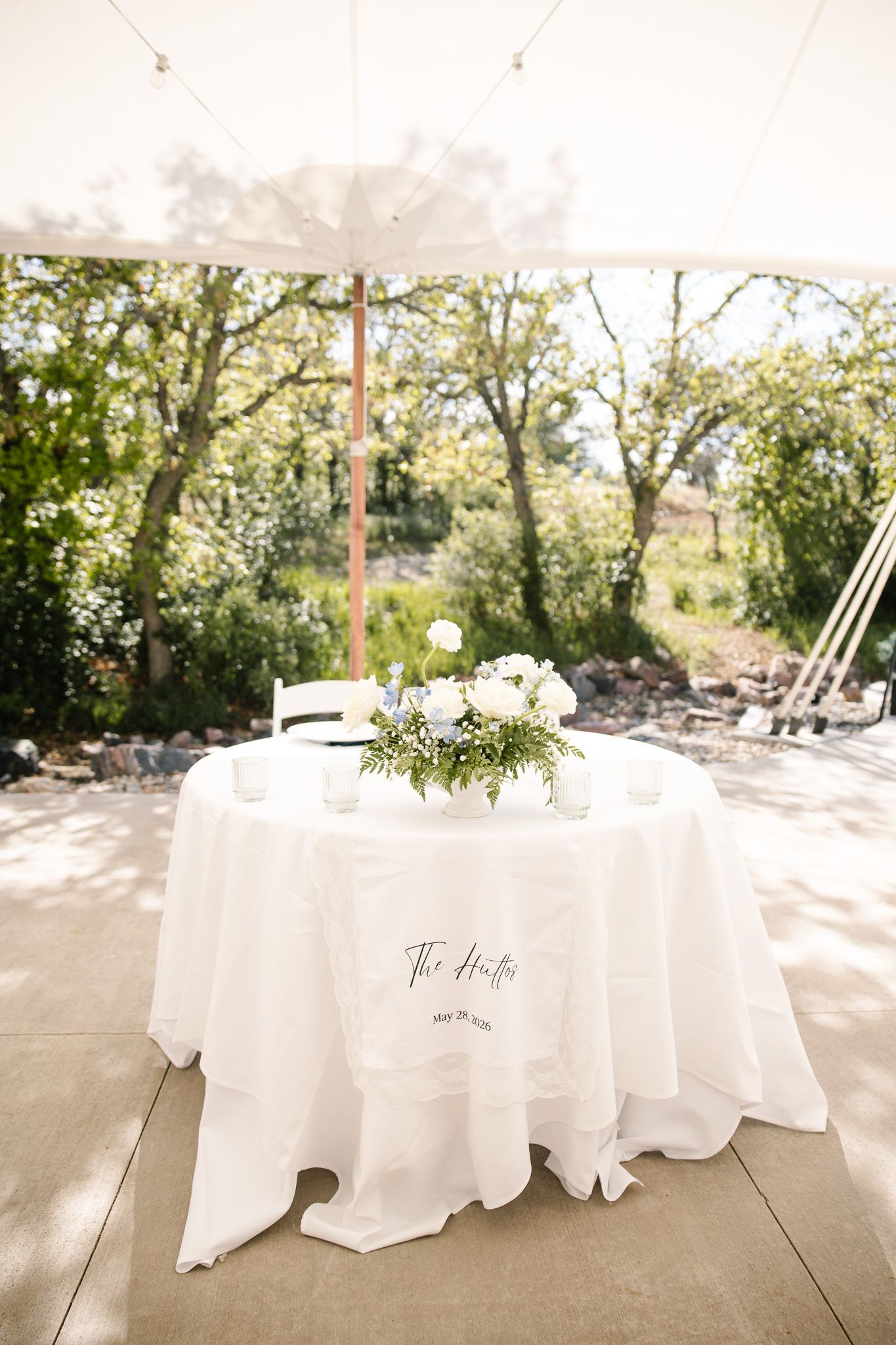 Elegant wedding sweetheart table with white linen, floral centerpiece, and personalized tablecloth under a tent outdoors.