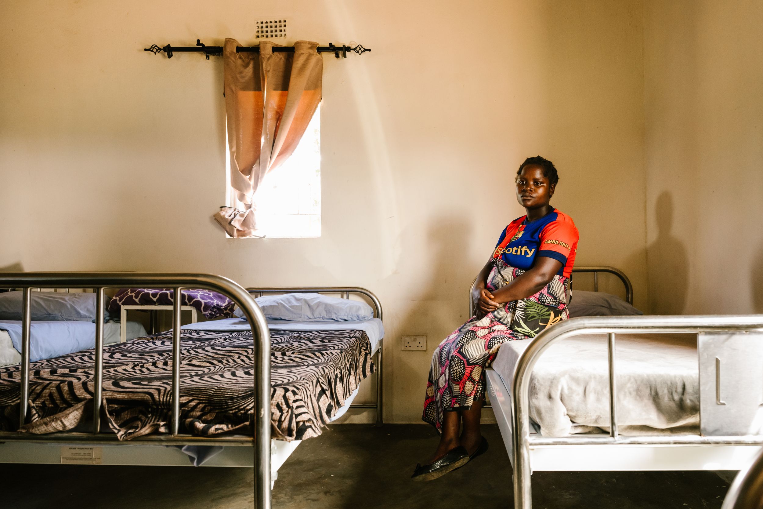 Woman sitting on a metal bed frame in a sparse room with two beds and a small window.