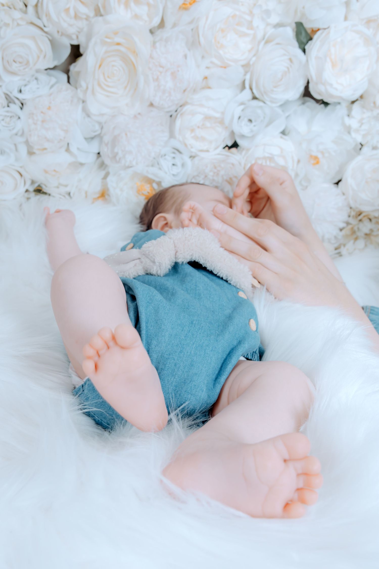 Newborn baby in blue onesie sleeping on white fur blanket surrounded by white roses with mother's hand nearby.