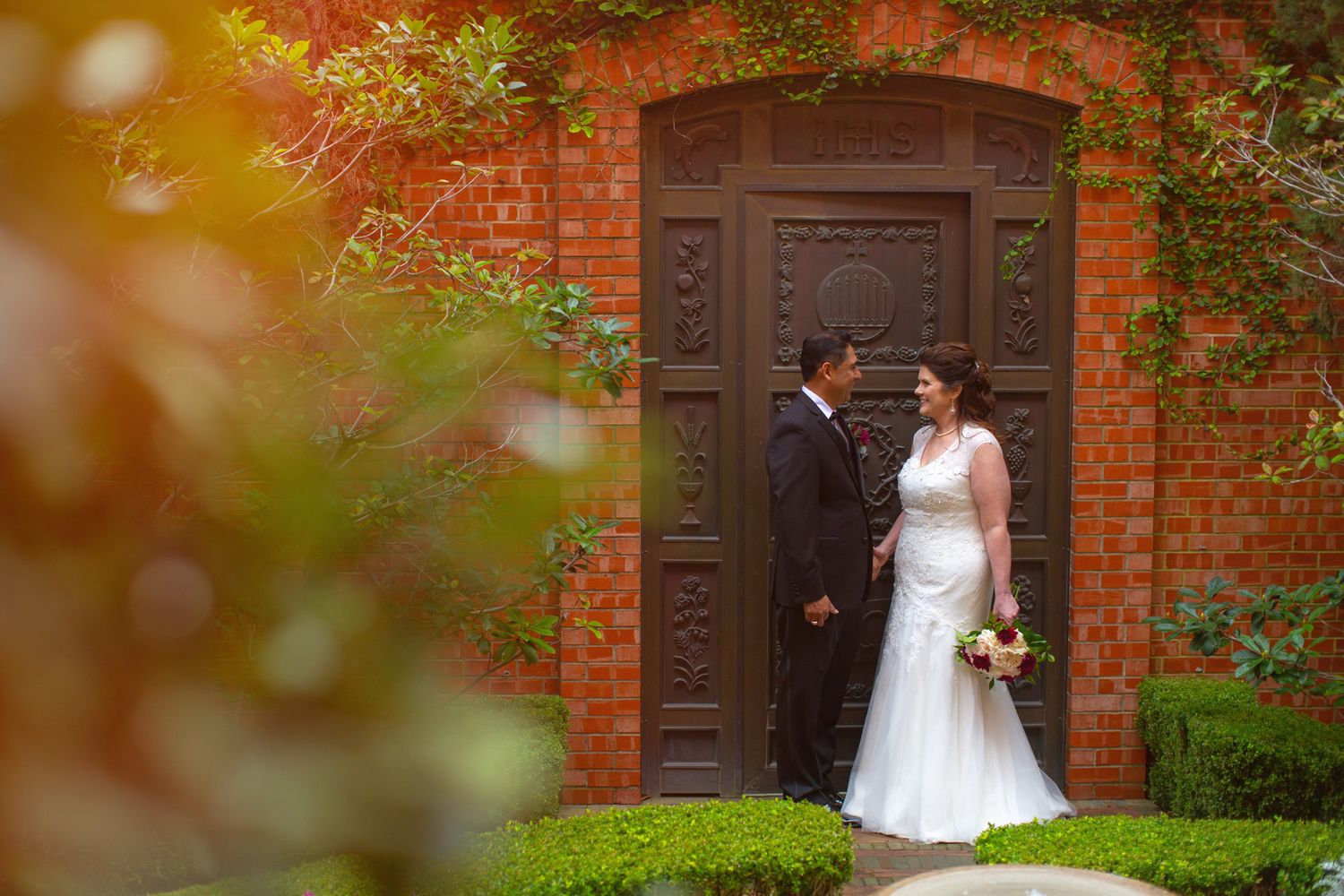 Bride and groom share a romantic moment before an ornate wooden door on a red brick wall surrounded by lush greenery.