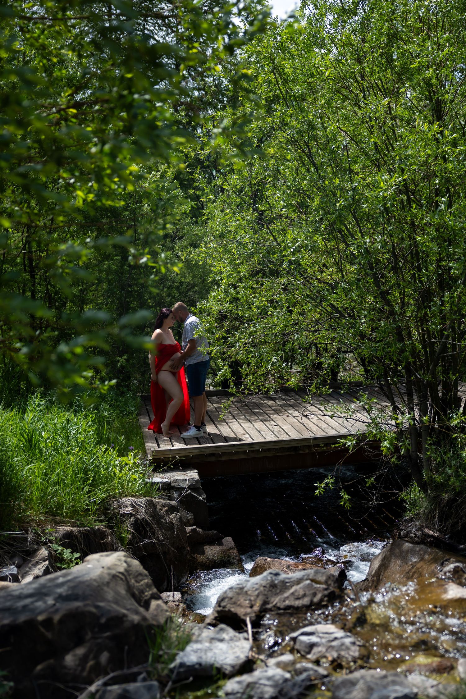 Couple embracing on a wooden bridge over a rocky stream surrounded by lush green trees in a forest.