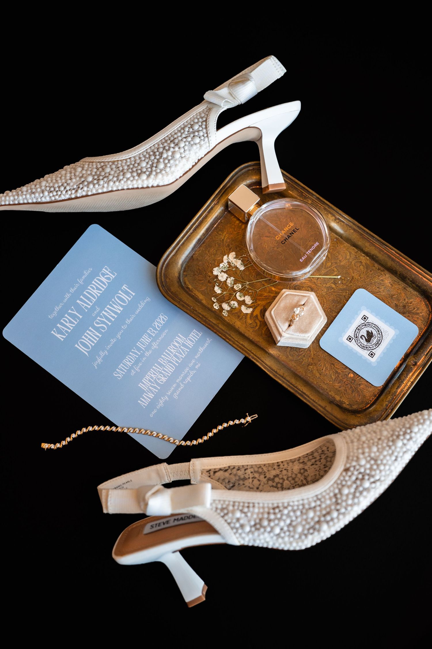 Pearl-embellished bridal heels, blue wedding invitation, gold tray with perfume, ring box, and earrings on black background.