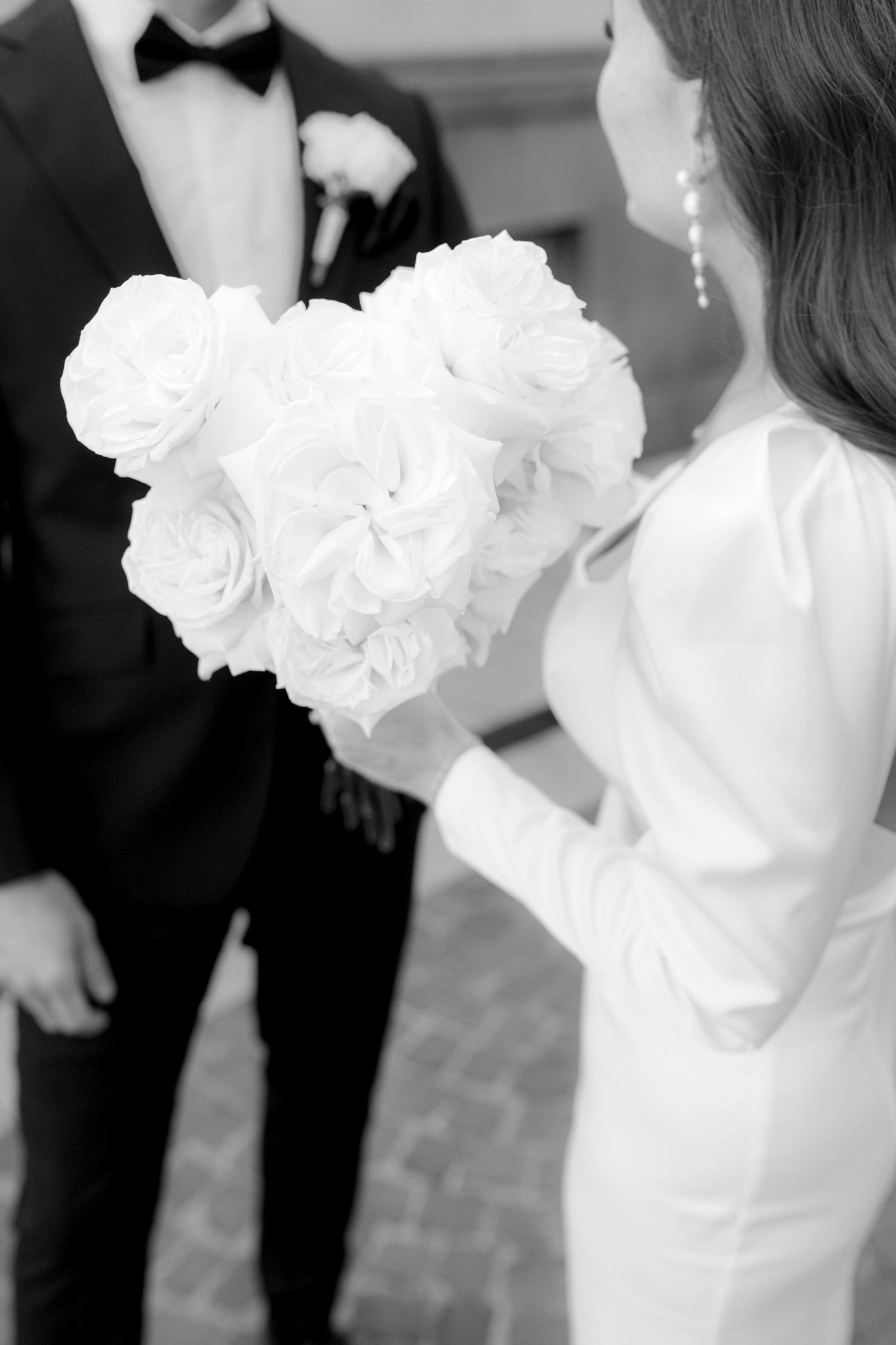 Black and white photo of bride holding white rose bouquet beside groom in tuxedo at elegant wedding ceremony.