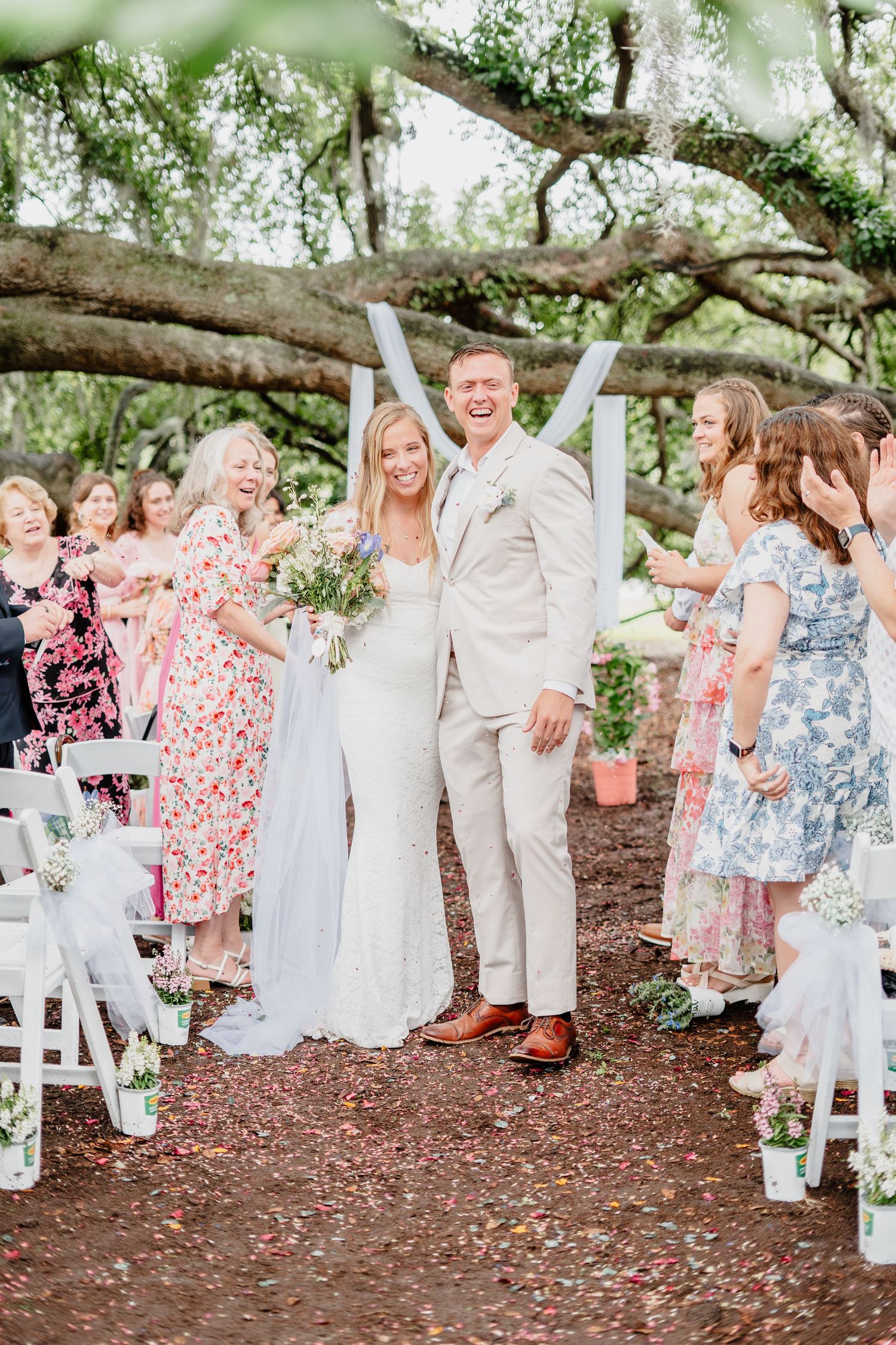 Bride and groom walk down outdoor aisle after wedding ceremony as guests celebrate with colorful confetti.