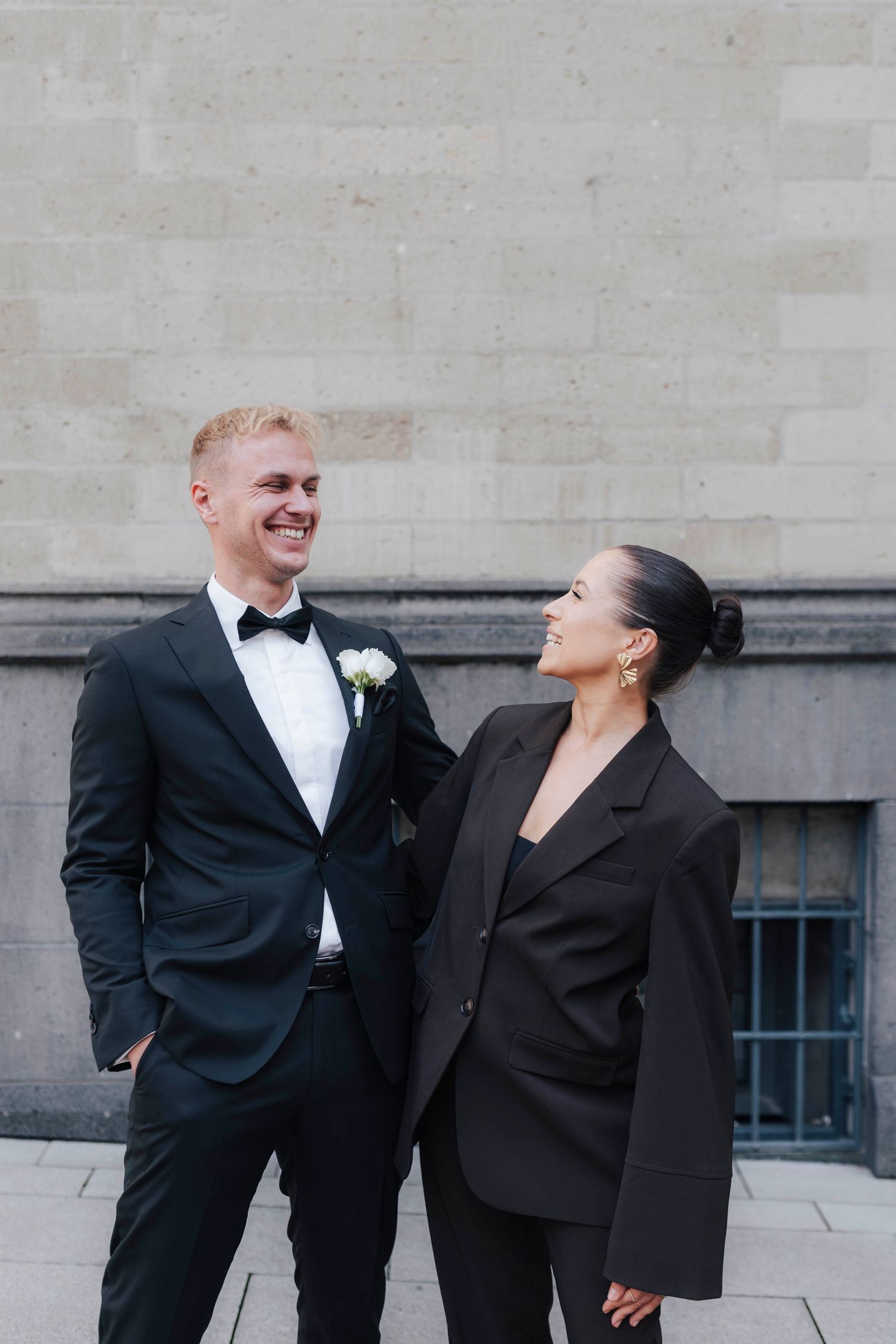 A smiling groom in a black tuxedo with boutonniere stands beside a woman in a black suit outside a stone building.