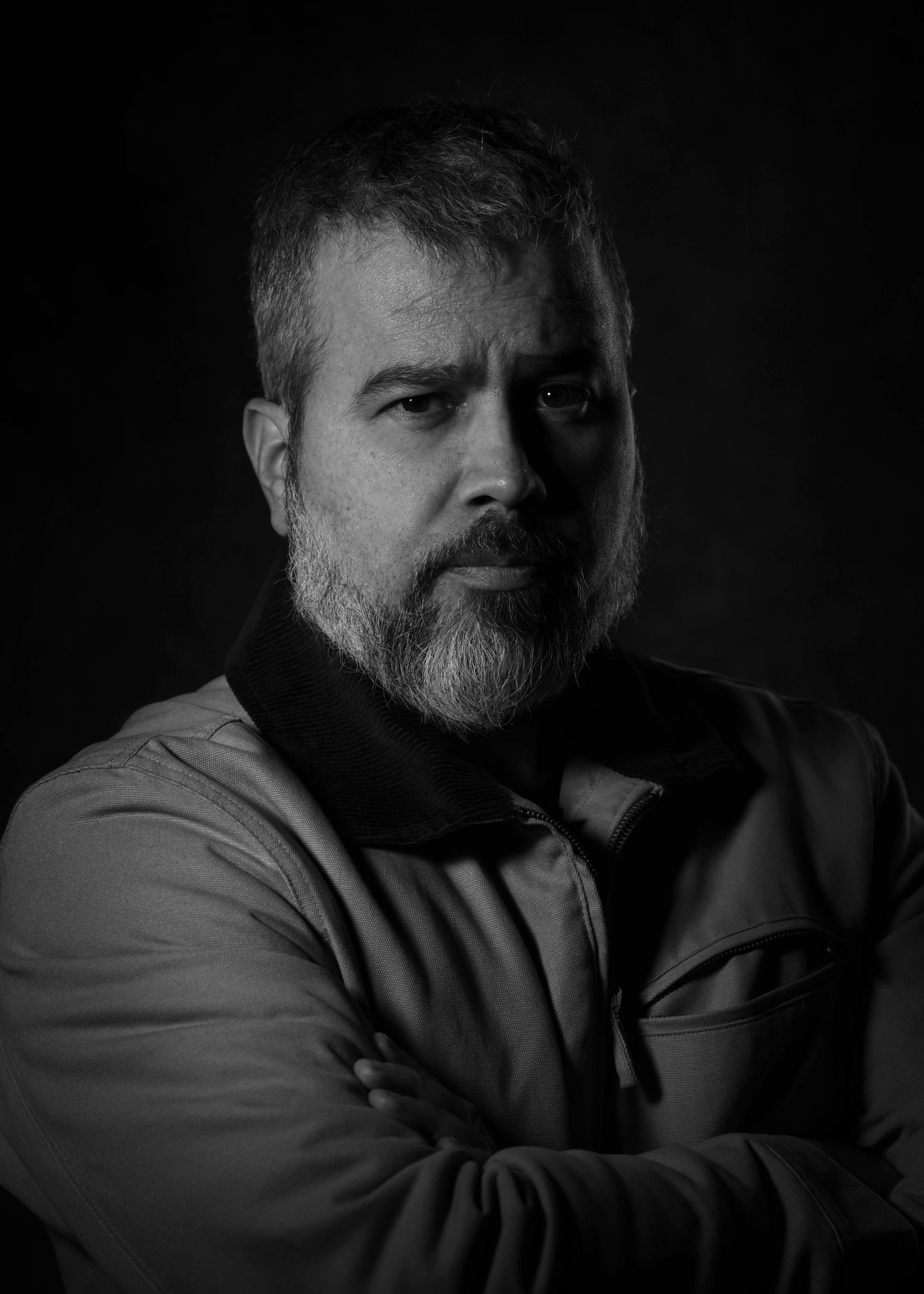 Black and white portrait of a middle-aged bearded man with arms crossed against a dark background.