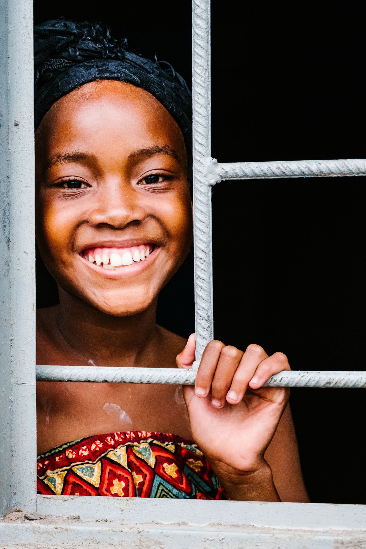 Smiling young girl peering through metal window bars, wearing colorful patterned top, joyful expression.