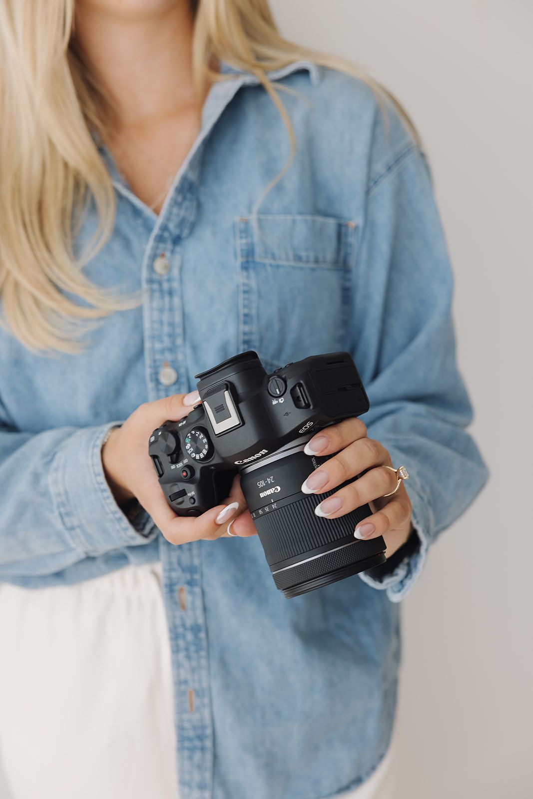 Woman in denim shirt holding a Canon DSLR camera with manicured nails and gold ring.