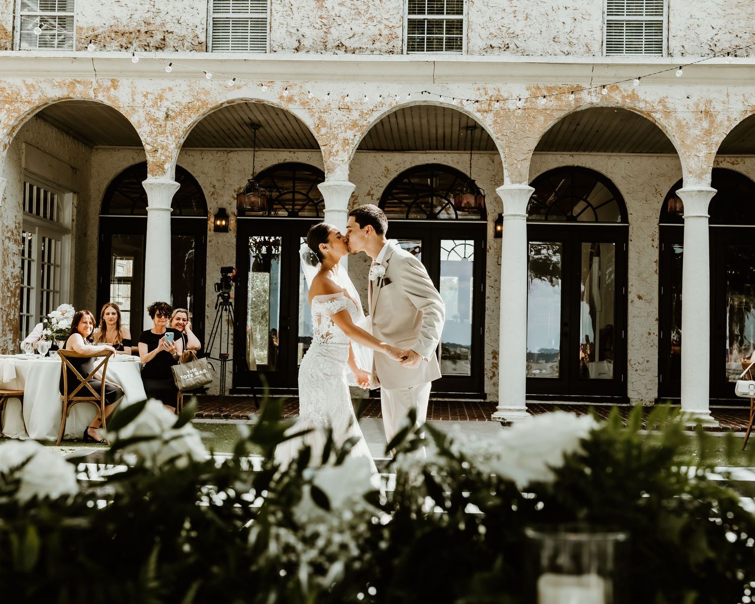 Elegant outdoor wedding reception in a courtyard with arched colonnade, lush greenery centerpieces, and couple dancing.