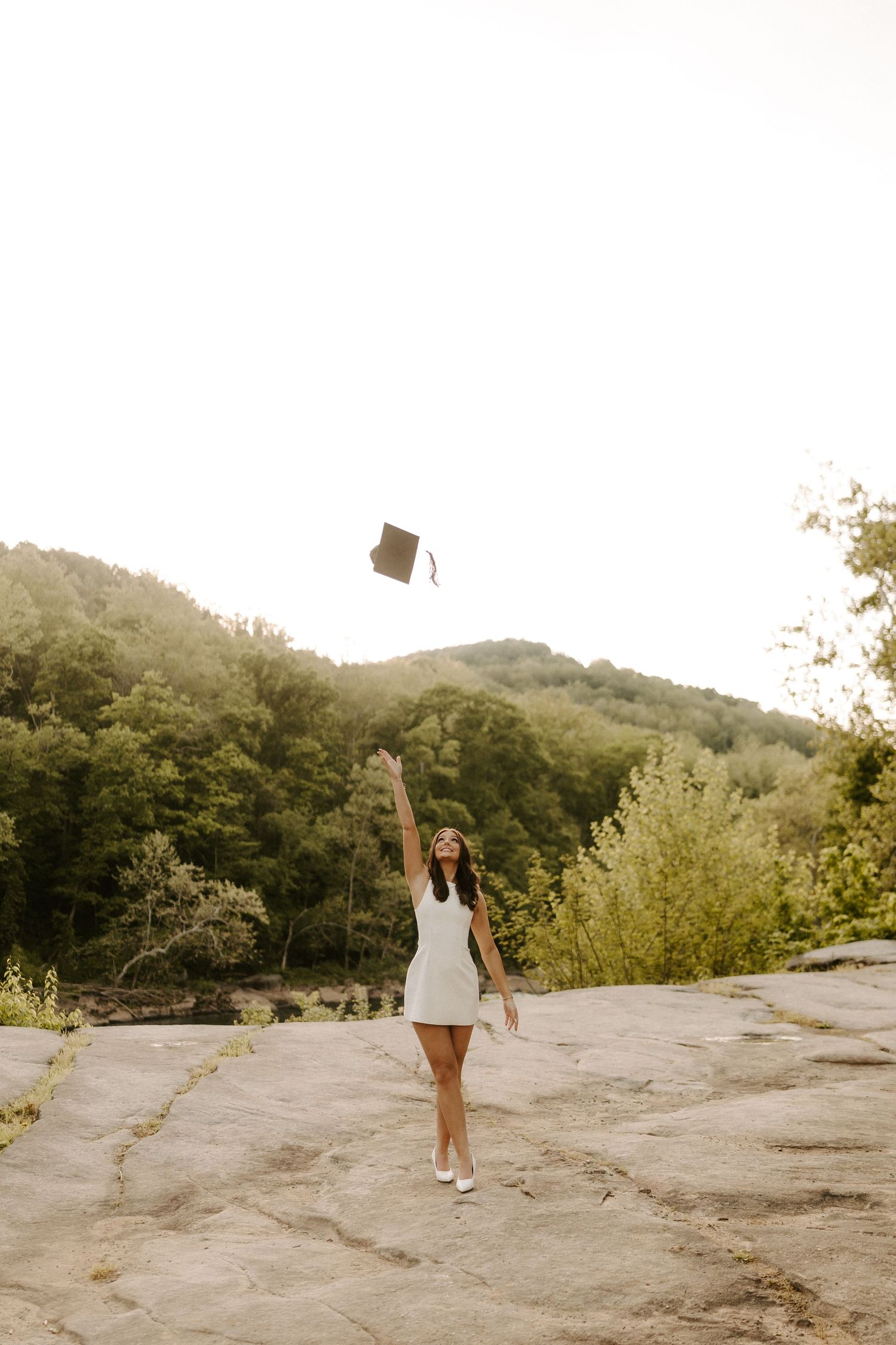 Young woman in white dress tossing graduation cap in the air on rocky overlook with lush green mountains behind her.