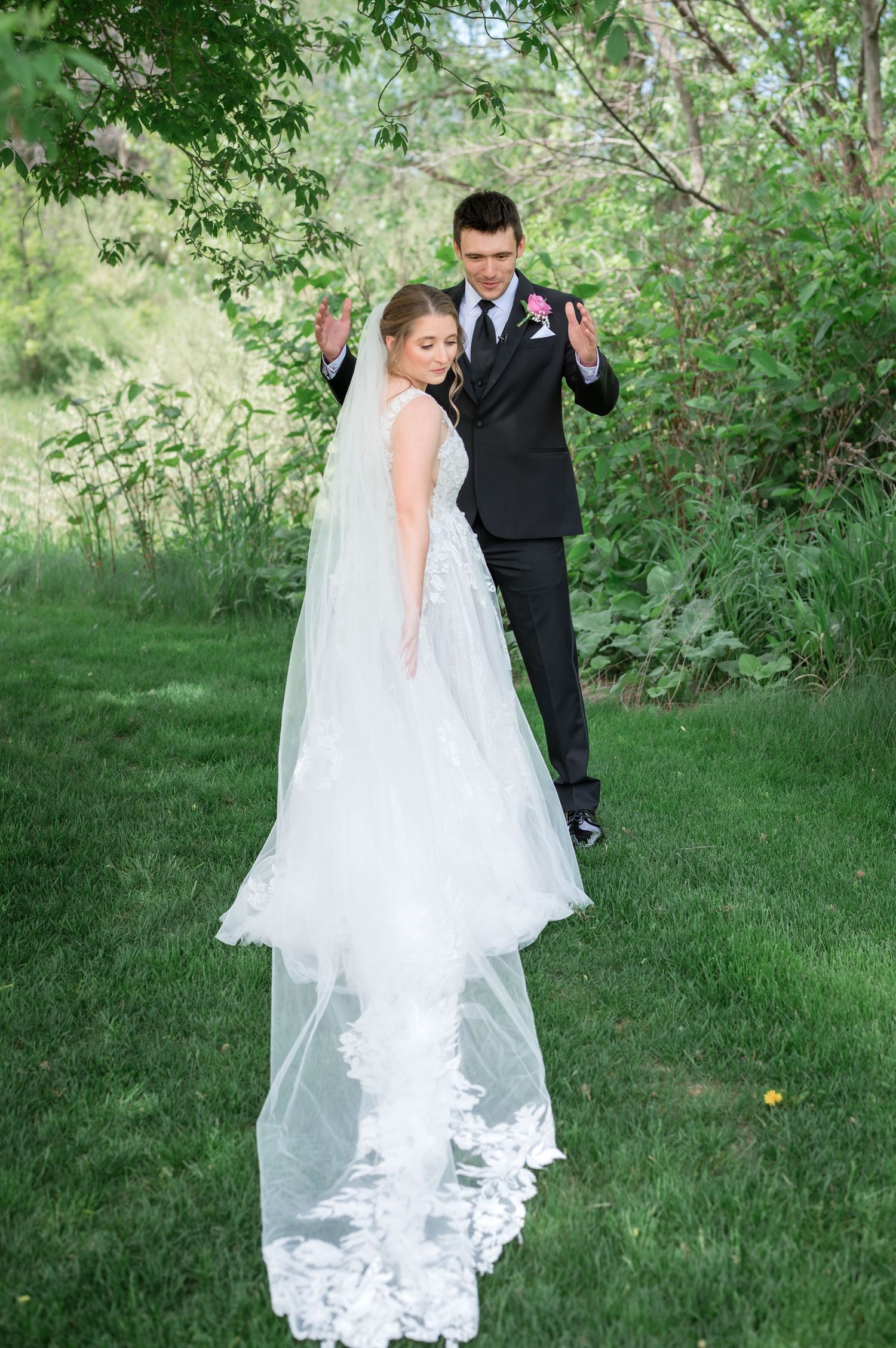 Bride in white gown with lace veil and groom in black suit share a joyful first look moment outdoors.