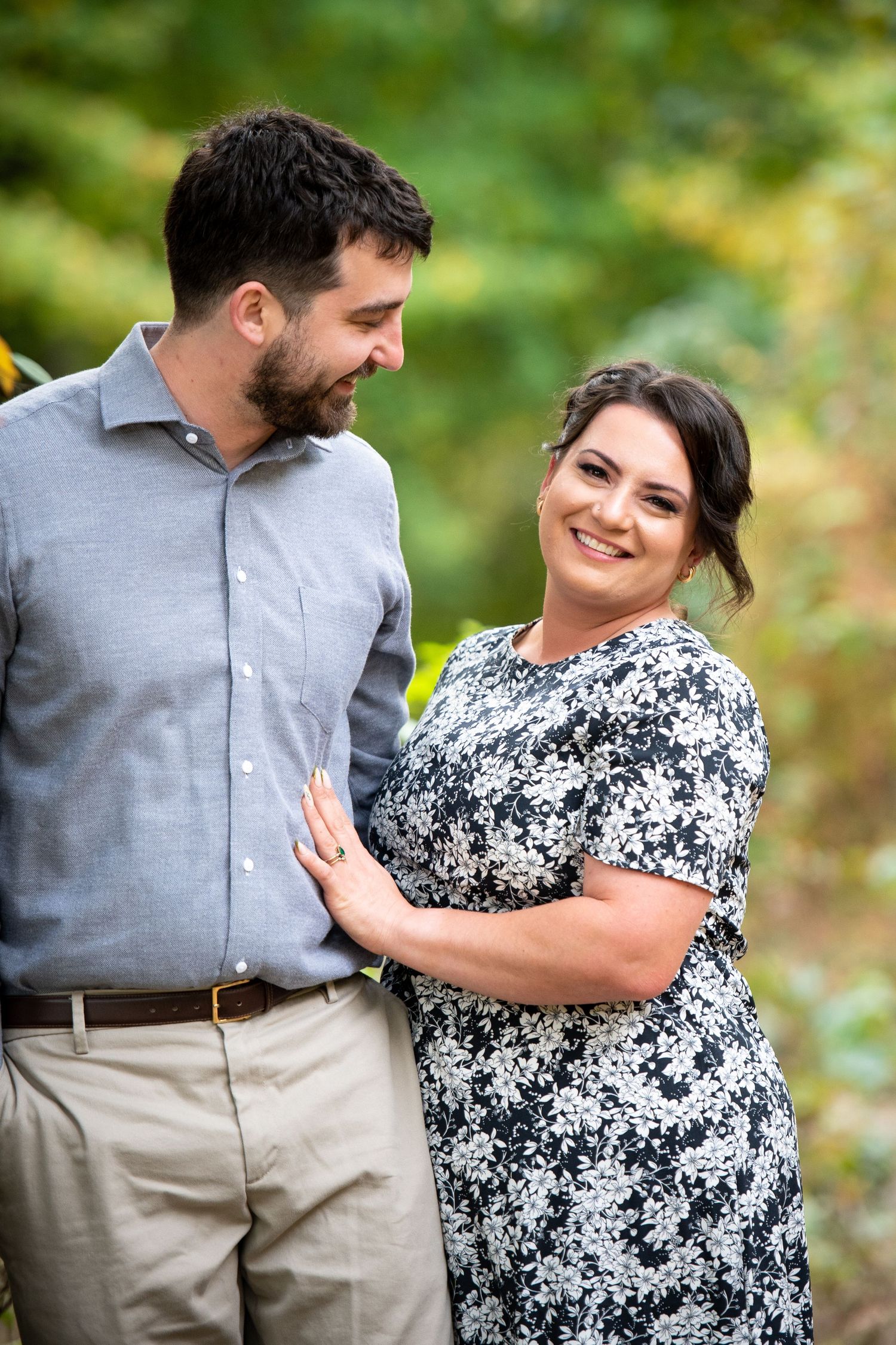 A smiling couple poses outdoors, woman in floral dress and man in gray shirt, surrounded by lush green foliage.