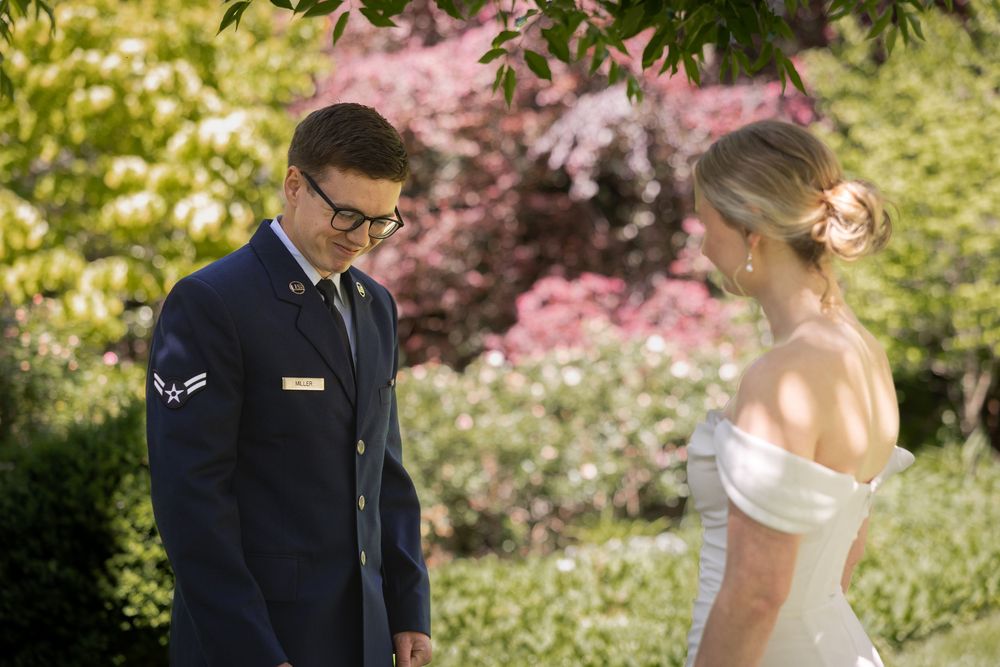 Military groom in dress uniform smiles at bride in off-shoulder wedding gown during outdoor first look ceremony.