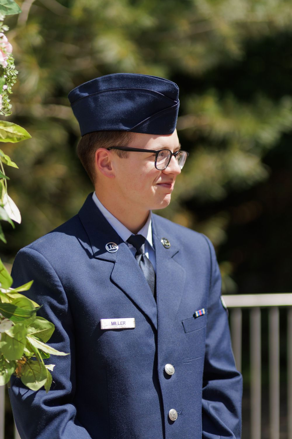 Young man in US Air Force dress uniform with cap and glasses stands outdoors at a formal military ceremony.