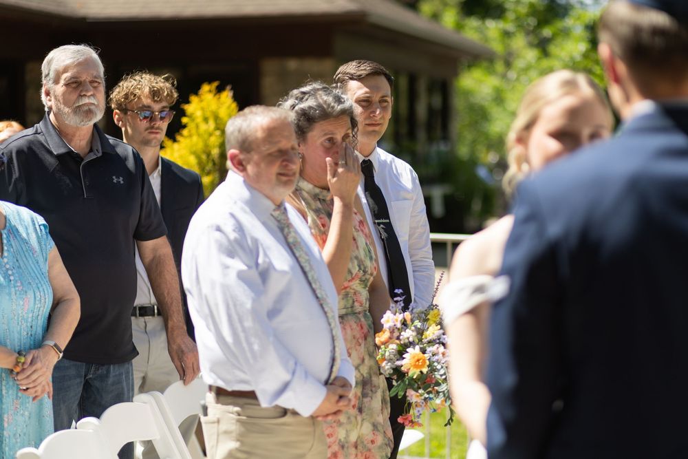 Emotional wedding guests watching outdoor ceremony, man in white shirt wiping tears, surrounded by family and friends.
