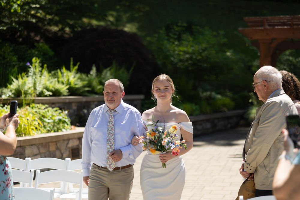Bride walks down outdoor aisle holding colorful bouquet, escorted by man in blue tie at sunny garden wedding ceremony.