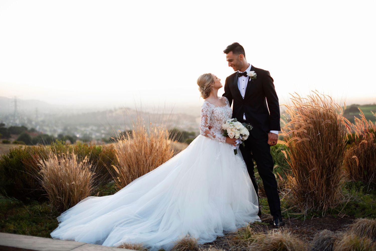 Bride in a lace-sleeved gown gazing up at her groom among tall grasses on a hillside at golden hour at Almaden Boulder Ridge Country Club.