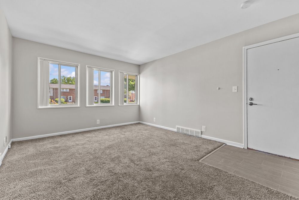 Bright empty living room with brown carpet, large windows, white door, and gray walls.