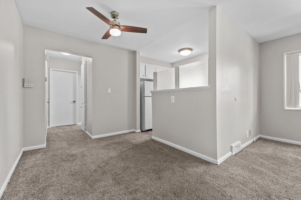 Bright empty living room with beige carpet, ceiling fan, white walls, and view into kitchen with refrigerator.