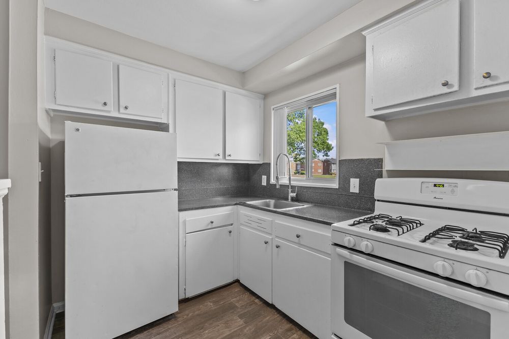 White kitchen with gas range, refrigerator, dark countertops, white cabinets, and a window above the sink.